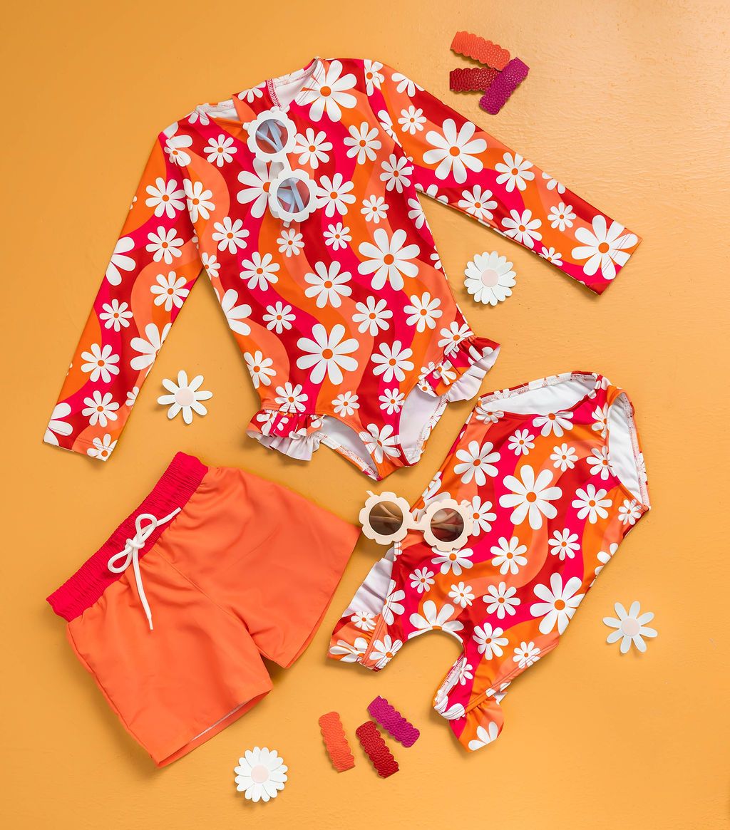flat lay image bonbon swim retro daisy swim suits with daisy flower hair clips, daisy sun glasses, boys orange and pink swim trunks on an orange background