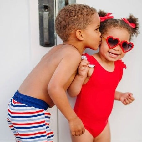 little boy kissing little girls cheek. Boy is wearing blue and red striped swim trunks and the girl is wearing a red one piece swimsuit